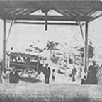 Black-and-white image of people around a vehicle under a wooden structure.