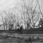 Black-and-white photo of a fence and leafless trees near houses.